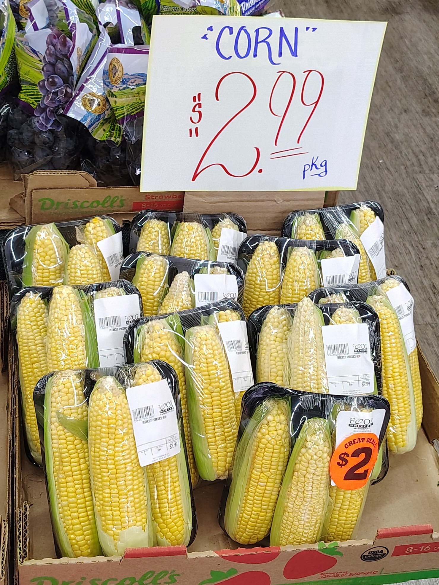 Display of packaged ears of corn at a grocery store. The display is labeled with a handwritten sign reading "corn $2.99 per package" but the word "corn" is in unnecessary quotation marks.