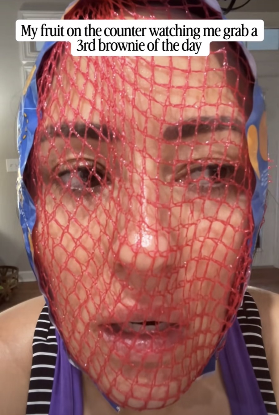 A person with their face inside a net commonly used for oranges with the caption, “My fruit on the counter watching me grab a 3rd brownie of the day”