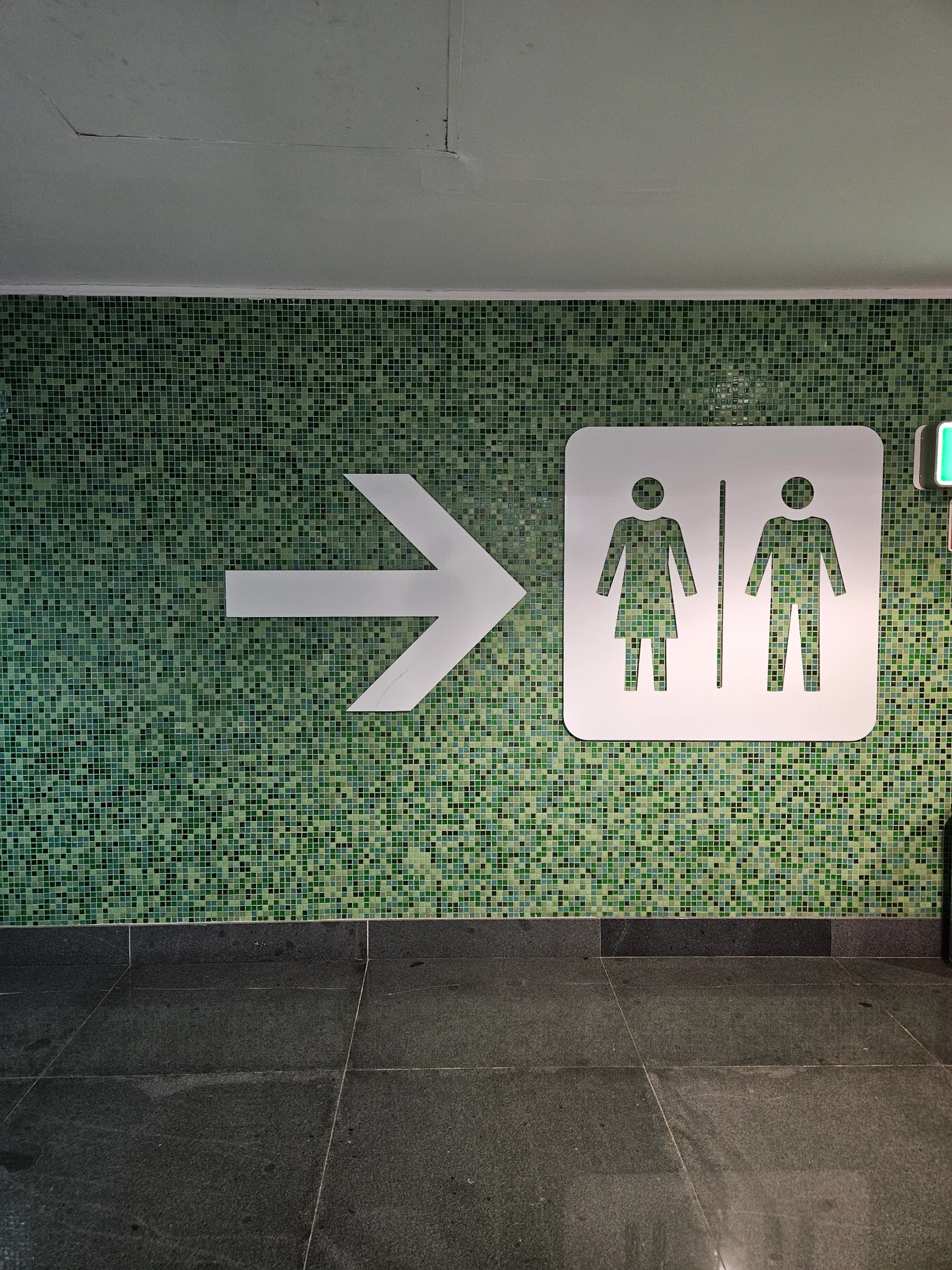 Wall with a bunch of green tiles in different hues. A sign leading to the bathroom